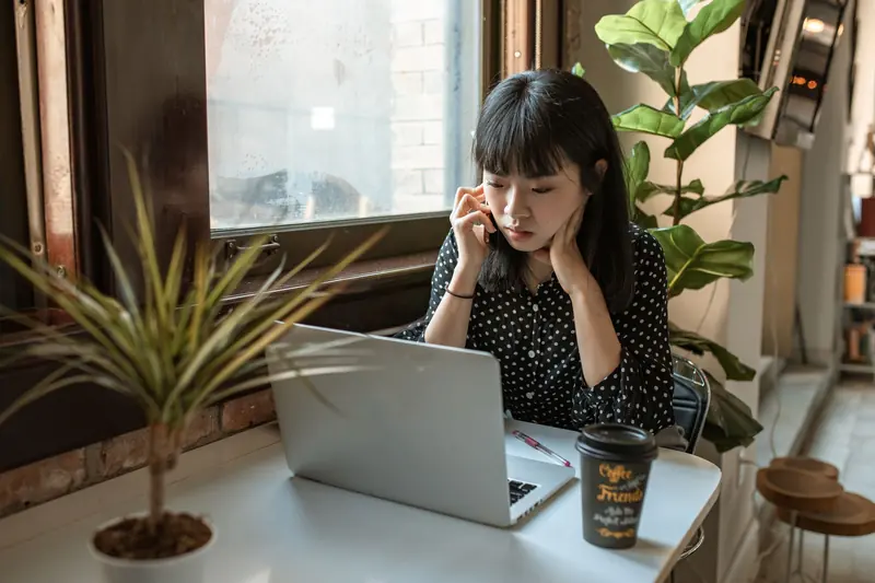 Asian woman taking a call while working on a laptop by the window